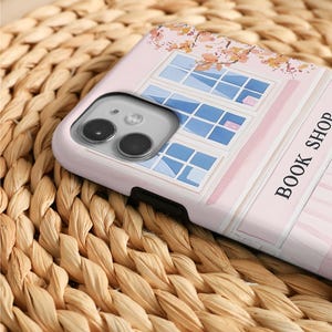 May include: A pink phone case featuring a watercolor illustration of a window with blue panes and a floral design. The words "BOOK SHOP" are printed vertically on the right side of the case. The case is on a woven surface.
