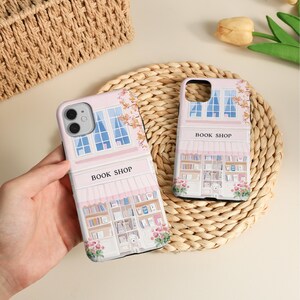 May include: Two pink phone cases with a book shop design. The cases feature a facade with windows, a striped awning, and shelves of books. The text "BOOK SHOP" is printed on the awning. Floral accents and a small white dog are also visible.