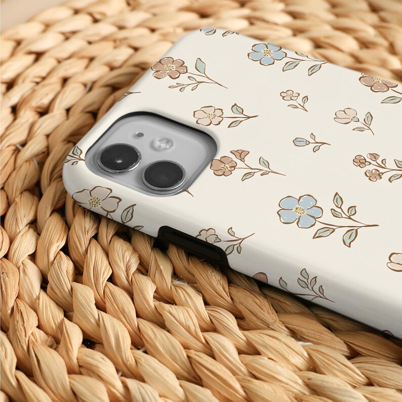 K&ouml;nnte beinhalten: Eine cremefarbene Smartphone-H&uuml;lle mit einem wiederkehrenden Muster aus kleinen, handgezeichneten Blumen in gedeckten Blau-, Braun- und Salbeigr&uuml;nt&ouml;nen. Die H&uuml;lle hat einen rechteckigen Ausschnitt f&uuml;r ein Dual-Kamera-System oben links.