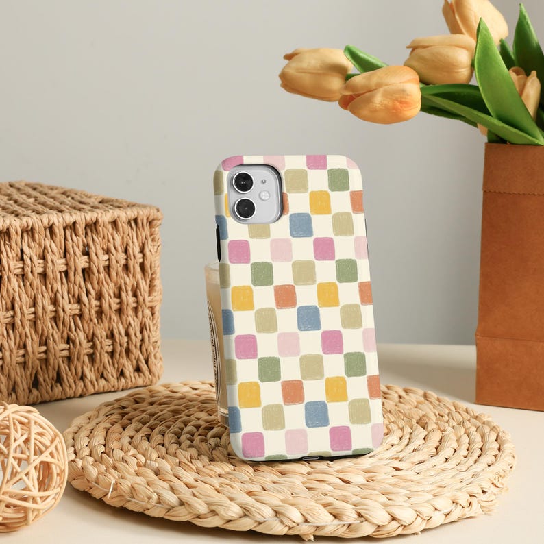 K&ouml;nnte beinhalten: Eine Smartphone-H&uuml;lle mit einem farbenfrohen Schachbrettmuster in Pastellt&ouml;nen von Rosa, Blau, Gelb und Gr&uuml;n. Die H&uuml;lle befindet sich vor einem neutralen Hintergrund mit einem Weidenkorb und Blumen.