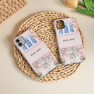 May include: Two pink phone cases with a book shop design. The cases feature a detailed illustration of a bookstore facade with windows, awnings, and the text "BOOK SHOP". Floral accents and a woven background complete the scene.