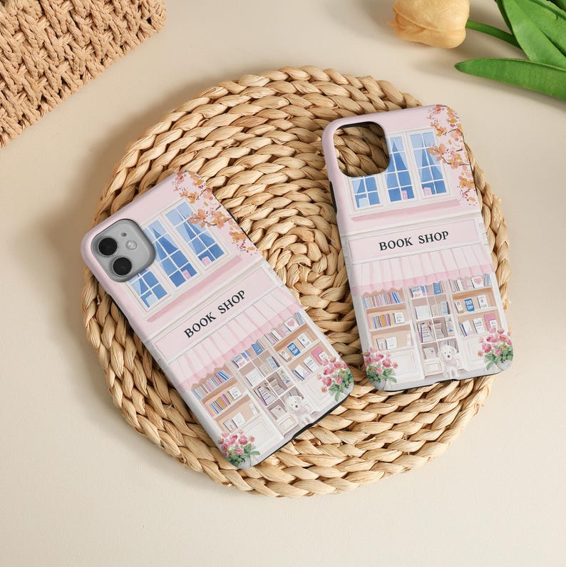 May include: Two pink phone cases with a book shop design. The cases feature a detailed illustration of a bookstore facade with windows, awnings, and the text "BOOK SHOP". Floral accents and a woven background complete the scene.