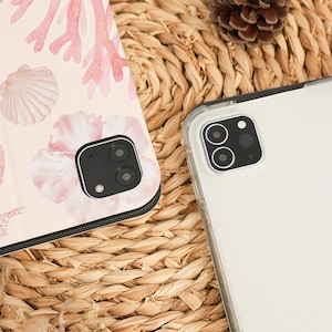 Pu&ograve; includere: Primo piano di due custodie per telefono su una superficie tessuta. Una custodia presenta un design di corallo rosa e conchiglie su uno sfondo chiaro. L'altra &egrave; una custodia trasparente. Una pigna &egrave; sullo sfondo.