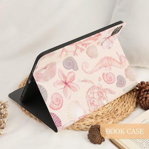 Pu&ograve; includere: Una custodia per tablet con un motivo ad acquerello rosa chiaro raffigurante creature marine e flora, tra cui una tartaruga marina, un cavalluccio marino, varie conchiglie, corallo e fiori tropicali. La custodia ha una cornice nera.