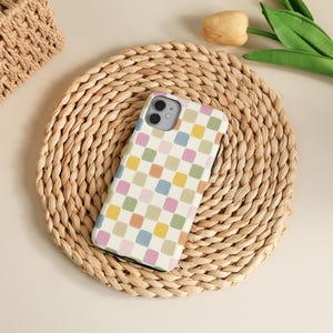 K&ouml;nnte beinhalten: Eine Smartphone-H&uuml;lle mit einem farbenfrohen Schachbrettmuster in Pastellt&ouml;nen von Rosa, Blau, Gelb und Gr&uuml;n. Die H&uuml;lle liegt auf einem runden, gewebten Platzset.