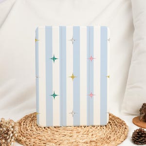 May include: A light blue and white striped tablet case with a pattern of colorful starbursts. The case is rectangular and has a folding cover. The case is sitting on a woven mat.