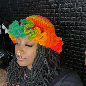 May include: A person wearing a rainbow crochet hat with a flower design. The hat is made of colorful yarn and has a unique, textured look.