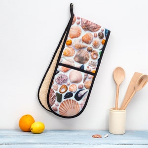 May include: A white oven mitt with a black trim and a colourful pattern of seashells. The mitt is hanging from a hook on a white wall.
