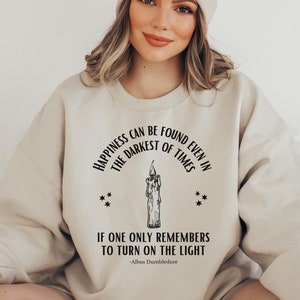 May include: A beige sweatshirt with a black graphic print of a candle and the text "Happiness can be found even in the darkest of times. If one only remembers to turn on the light - Albus Dumbledore."