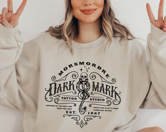 May include: A beige sweatshirt with a black graphic design of a skull and snake with the text "MORS MORDRE DARK MARK TATTOO STUDIO APPROVED BY THE DARK LORD 500 PER MARK EST. 1967 KNOCHTURN ALLEY'S FINEST PUREBLOOD ARTIS"