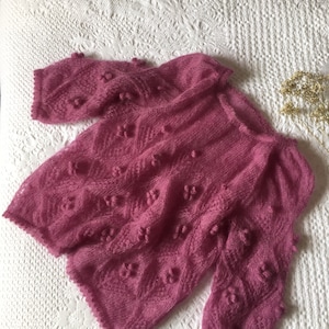May include: A pink knitted sweater with a delicate openwork pattern and small pom-poms throughout.
