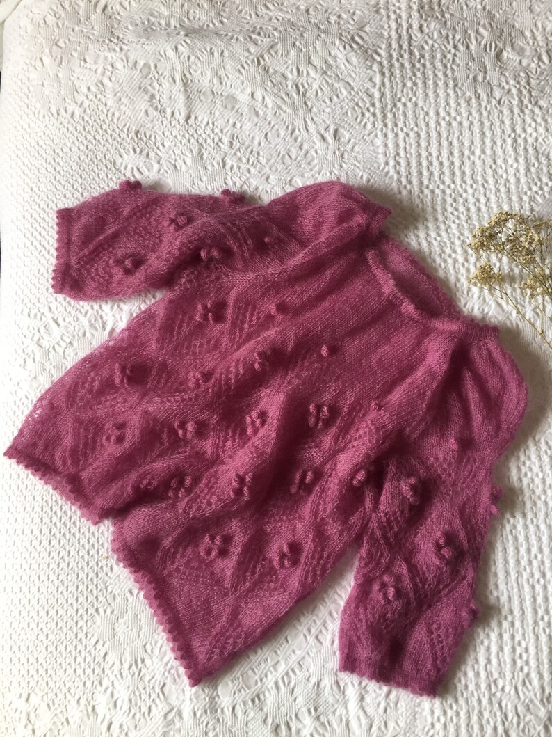 May include: A pink knitted sweater with a delicate openwork pattern and small pom-poms throughout.
