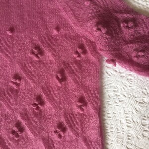 May include: A pink knitted sweater with a delicate, lacy pattern and small, raised bobbles throughout. The sweater has a loose, flowing fit and a soft, airy texture.