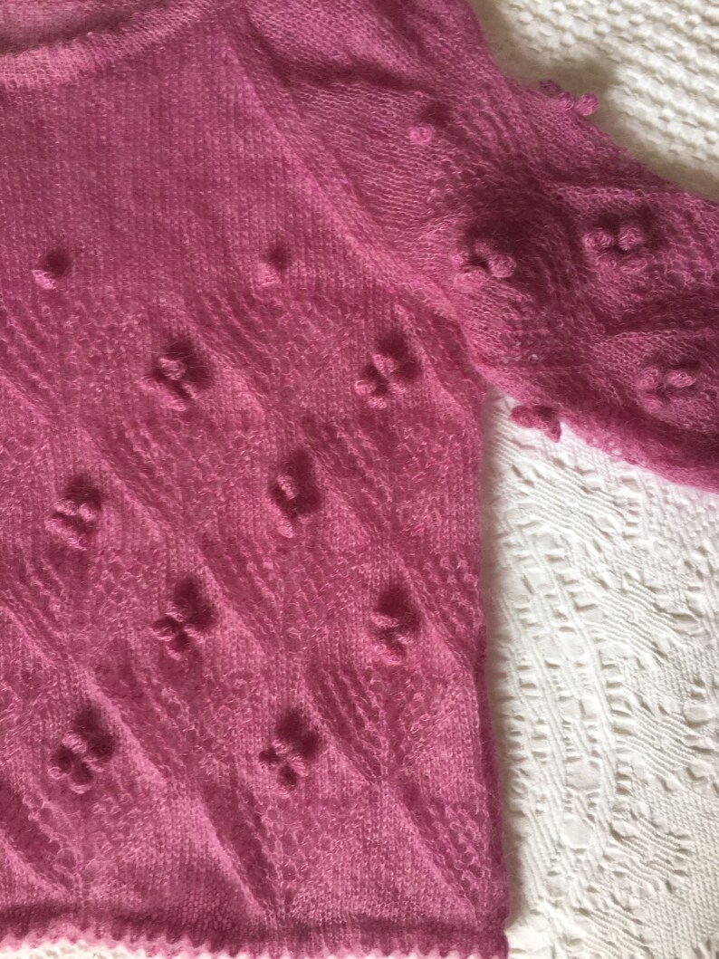 May include: A pink knitted sweater with a delicate, lacy pattern and small, raised bobbles throughout. The sweater has a loose, flowing fit and a soft, airy texture.