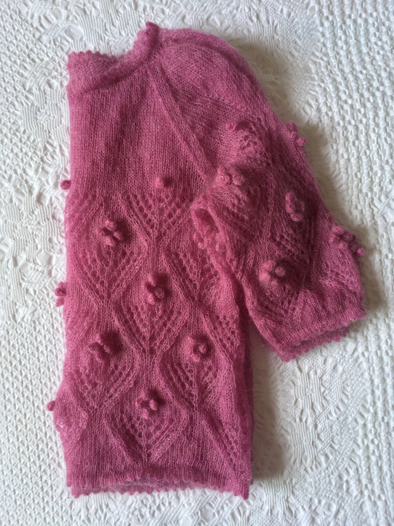 May include: A pink knitted sweater with a delicate lace pattern and small, raised bobbles. The sweater has a slightly loose fit and a round neckline.