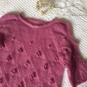 May include: A pink knitted sweater with a textured pattern and small pom-poms throughout.
