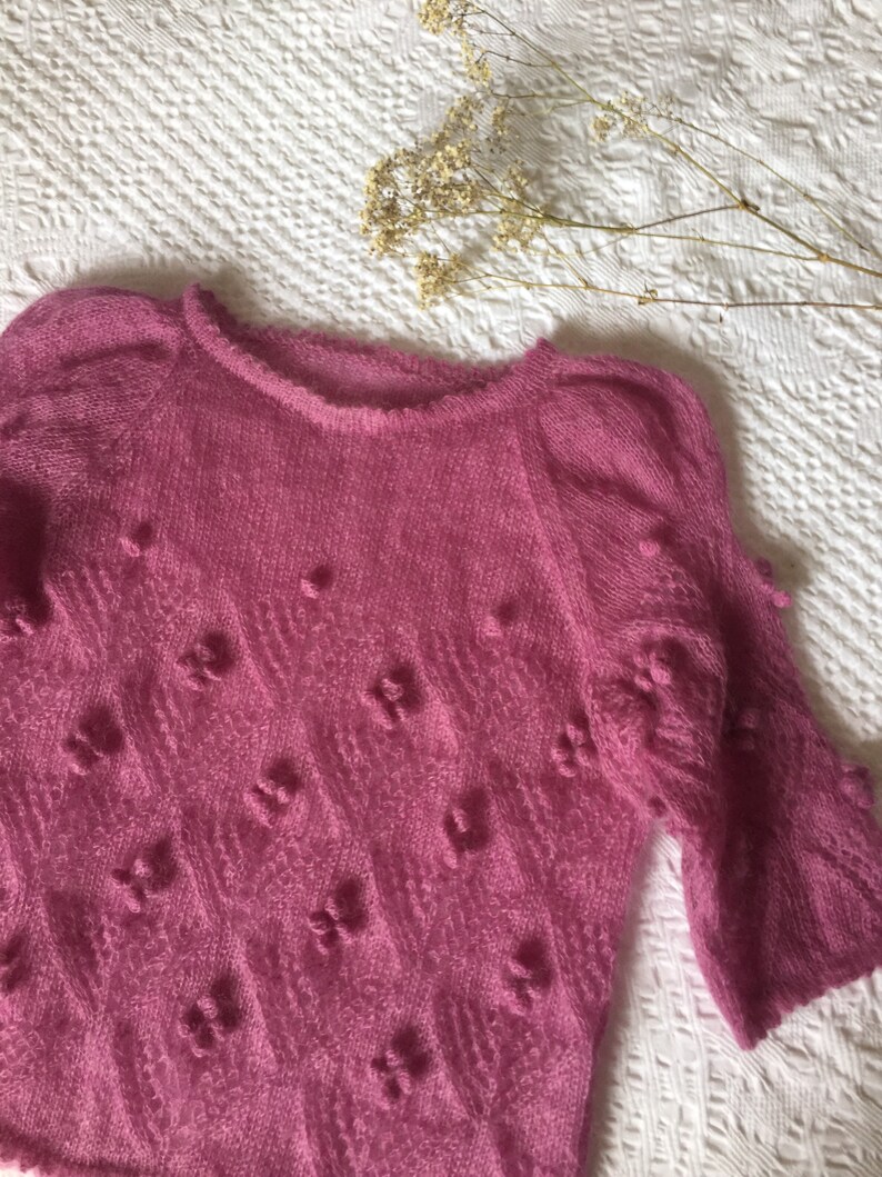 May include: A pink knitted sweater with a textured pattern and small pom-poms throughout.