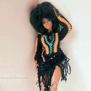 Peut inclure: Une poupée noire avec une coiffure afro portant une tenue crochetée noire, orange et verte avec des franges. La poupée est debout devant un mur rose clair. Le texte "SUNSET IN DOLLCITY" est visible en bas de l'image.