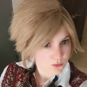 Könnte beinhalten: Eine Person in einem Steampunk-inspirierten Outfit. Das Outfit umfasst eine rote Weste mit goldenem Blumenmuster, braune Lederriemen und eine Brille. Die Person hat kurze, hellblonde Haare.