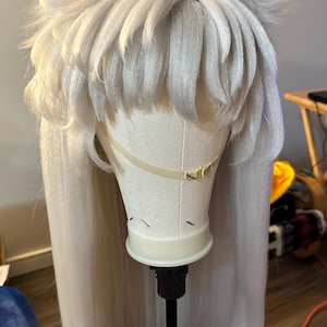 Puede incluir: Una peluca blanca con el pelo largo y liso y dos orejas de gato blancas y peludas. La peluca está en un soporte de peluca blanco.