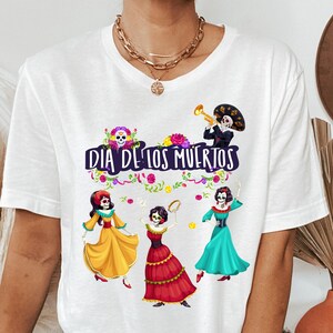 Op de afbeelding: Een wit t-shirt met een kleurrijke afbeelding van vier skeletfiguren in traditionele kleding, met de tekst "DIA DE LOS MUERTOS" in het paars gedrukt. Het ontwerp bevat bloemaccenten en een skelet dat een trompet bespeelt.