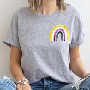 May include: A heather gray short-sleeve t-shirt featuring a small, watercolor-style rainbow graphic on the left chest. The rainbow design consists of concentric arches in yellow, white, purple, and dark green, with a textured, painted appearance.