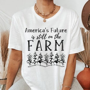 May include: White t-shirt with black text that reads "America's Future is still on the FARM" with a row of corn stalks below the text.