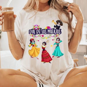 Op de afbeelding: Een cr&egrave;mekleurig t-shirt met een afbeelding van drie skeletfiguren in kleurrijke jurken en &eacute;&eacute;n skelet dat trompet speelt. De tekst "DIA DE LOS MUERTOS" is in het paars boven de figuren gedrukt, omringd door bloemmotieven.