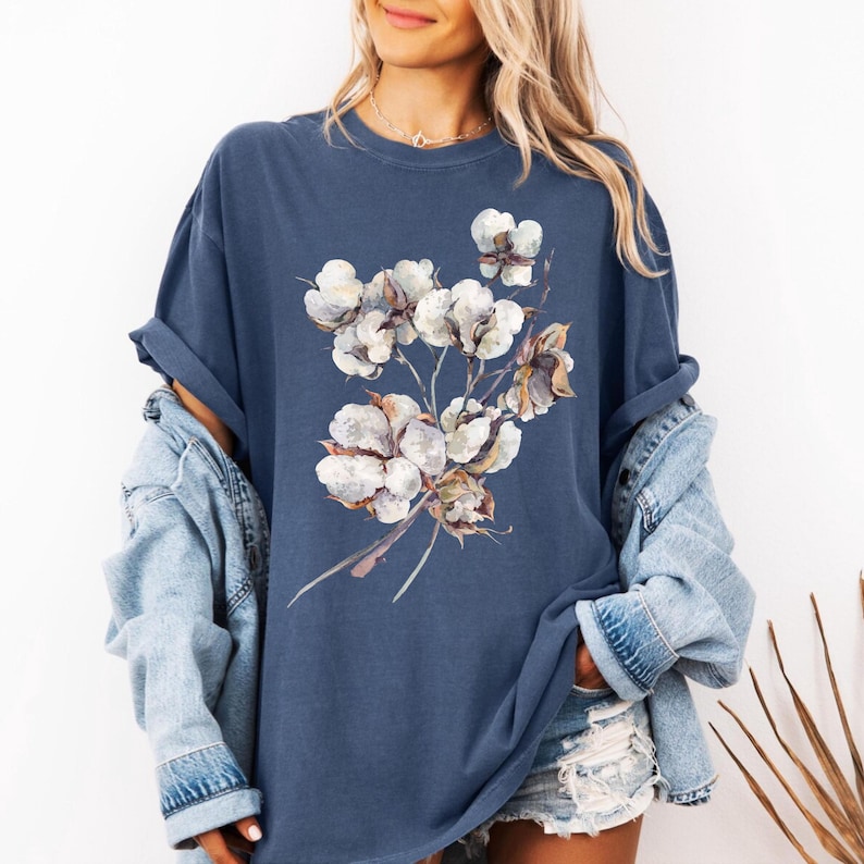 May include: A slate blue short-sleeve t-shirt featuring a central graphic print of a cotton plant branch with white, fluffy cotton bolls and brown stems. The shirt has a crew neckline and is layered under a light blue denim jacket.