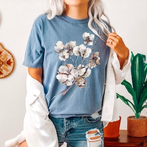 May include: A blue short-sleeved t-shirt featuring a central graphic print of white cotton bolls on brown stems. The shirt has a crew neckline and a relaxed fit, worn with a white corduroy button-down shirt layered over the shoulders.