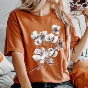 May include: A burnt orange short-sleeve t-shirt featuring a central graphic print of a cluster of white cotton bolls with brown stems and leaves. The shirt has a crew neckline and a relaxed fit, showcasing a detailed botanical illustration.