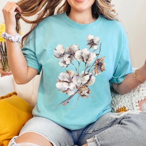 May include: A light blue short-sleeved t-shirt featuring a central graphic print of a cluster of white cotton bolls with brown stems and husks. The shirt has a crew neckline and a relaxed fit, showcasing a detailed botanical illustration.