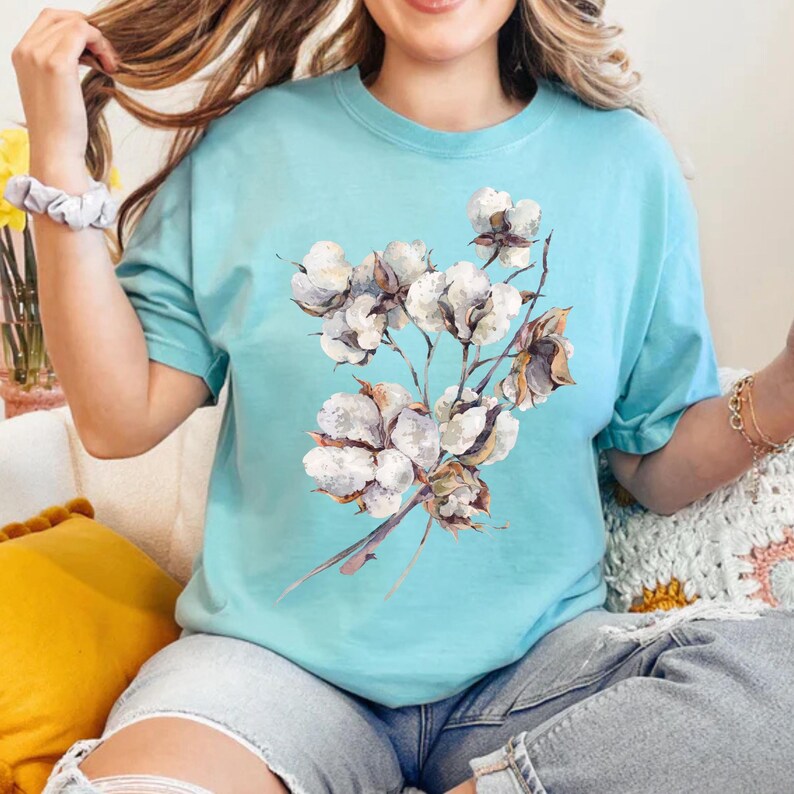 May include: A light blue short-sleeved t-shirt featuring a central graphic print of a cluster of white cotton bolls with brown stems and husks. The shirt has a crew neckline and a relaxed fit, showcasing a detailed botanical illustration.