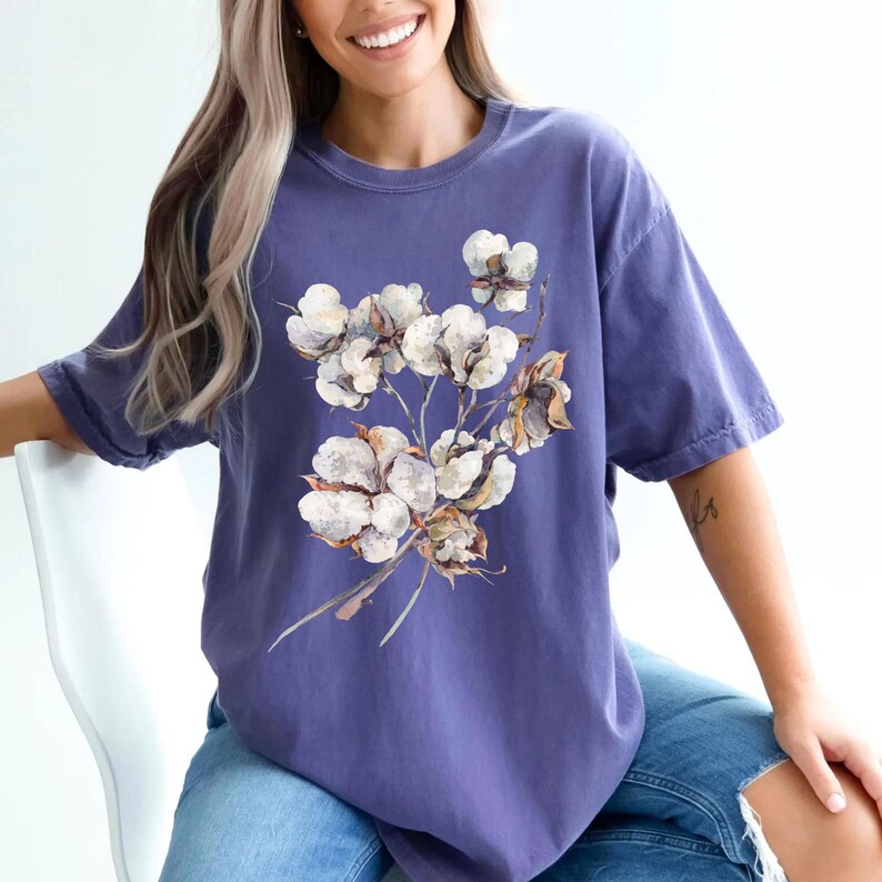 May include: A periwinkle blue short-sleeved t-shirt featuring a central graphic print of a cluster of white cotton bolls on brown stems. The shirt has a crew neckline and a relaxed fit, showcasing a detailed botanical illustration of cotton.
