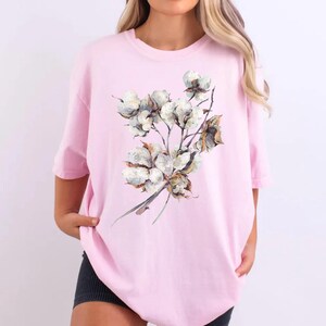 May include: A light pink short-sleeved t-shirt featuring a central graphic illustration of a cluster of white cotton bolls on brown stems. The design displays detailed textures of the fluffy white cotton and the woody branches against the fabric.