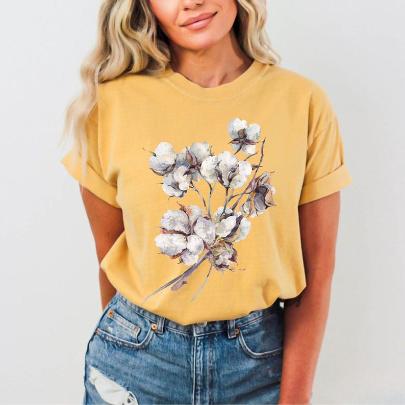 May include: A mustard yellow short-sleeve t-shirt featuring a central graphic illustration of a cluster of white cotton bolls with brown stems and husks. The shirt has a crew neckline and is paired with blue denim jeans with a distressed hem.