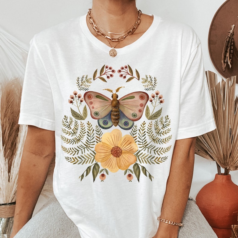 Pu&ograve; includere: T-shirt bianco con un'illustrazione ad acquerello di una farfalla con ali rosa, blu e marroni, circondata da una ghirlanda floreale. Un grande fiore giallo &egrave; in basso. La maglietta ha maniche corte e scollo tondo.