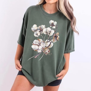 May include: An olive green short-sleeved t-shirt featuring a central graphic print of a cluster of white cotton bolls with brown stems and husks. The shirt has a crew neckline and a relaxed fit, showcasing a detailed botanical illustration.