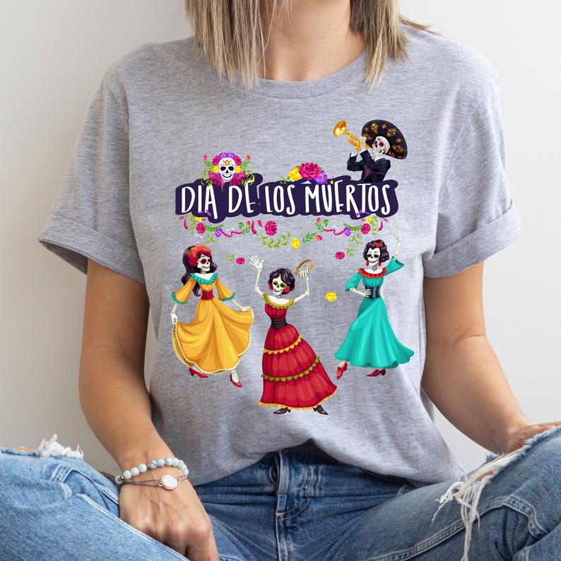 Op de afbeelding: Een grijs gem&ecirc;leerd t-shirt met een grafische print met de tekst DIA DE LOS MUERTOS. Het ontwerp bevat drie skeletten in kleurrijke jurken, een skelet dat trompet speelt, bloemaccenten en een schedel met bloemen aan de bovenkant.