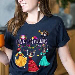 Op de afbeelding: Een donker marineblauw t-shirt met een grafische print met de tekst "DIA DE LOS MUERTOS". Het ontwerp bevat drie skeletfiguren in kleurrijke jurken, een skelet dat trompet speelt en bloemaccenten in roze, geel en groen.