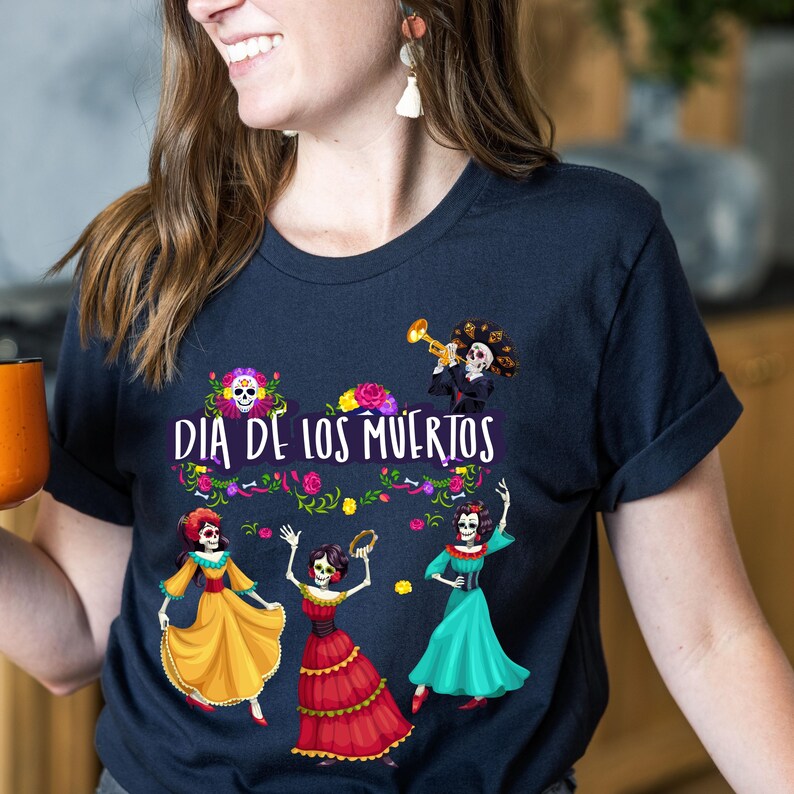 Op de afbeelding: Een donker marineblauw t-shirt met een grafische print met de tekst "DIA DE LOS MUERTOS". Het ontwerp bevat drie skeletfiguren in kleurrijke jurken, een skelet dat trompet speelt en bloemaccenten in roze, geel en groen.