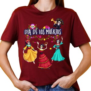 Op de afbeelding: Een bordeauxrood T-shirt met korte mouwen en een grafische print met de tekst "DIA DE LOS MUERTOS". Het ontwerp bevat drie skeletfiguren in kleurrijke jurken, een skelet dat trompet speelt en bloemaccenten in geel, roze en groen.