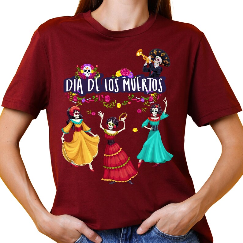 Op de afbeelding: Een bordeauxrood T-shirt met korte mouwen en een grafische print met de tekst "DIA DE LOS MUERTOS". Het ontwerp bevat drie skeletfiguren in kleurrijke jurken, een skelet dat trompet speelt en bloemaccenten in geel, roze en groen.