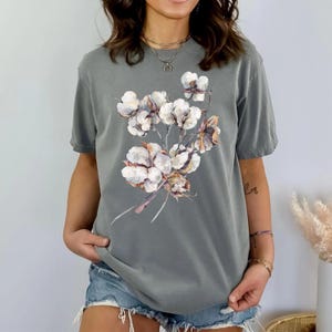 May include: A slate grey short-sleeve t-shirt featuring a central graphic print of a cluster of white cotton bolls with brown stems and soft, textured petals. The shirt has a crew neckline and a relaxed fit, showcasing a botanical illustration.