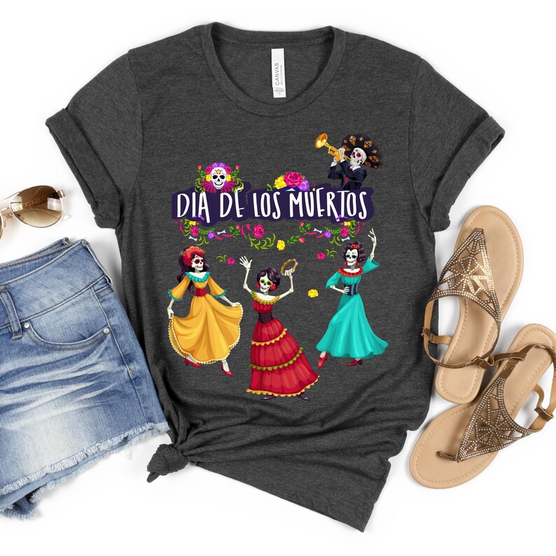 Op de afbeelding: Een donkergrijs t-shirt met korte mouwen en een afbeelding van vier skeletfiguren in kleurrijke jurken en een pak, met de witte tekst "DIA DE LOS MUERTOS", omringd door bloemaccenten en een trompettist bovenaan het ontwerp.