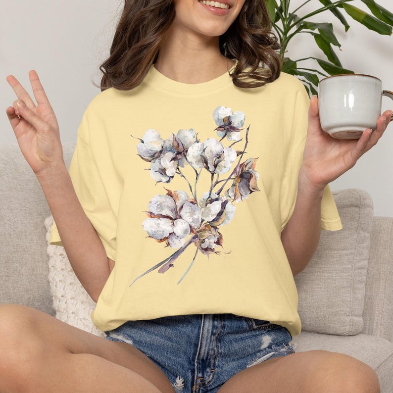 May include: A pale yellow short-sleeved t-shirt featuring a central watercolour-style illustration of a cotton plant branch with white fluffy bolls and brown stems. The shirt has a crew neckline and a relaxed fit, showcasing a botanical design.