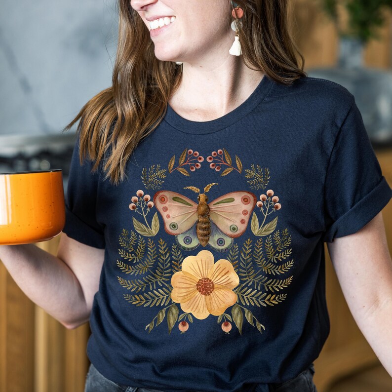 Pu&ograve; includere: T-shirt blu navy con un disegno di farfalla, un fiore giallo e dettagli botanici. La farfalla ha ali color camoscio con accenti rosa e blu. Il disegno &egrave; centrato sulla parte anteriore della maglietta.
