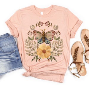 Pu&ograve; includere: Una maglietta rosa chiaro con un disegno di farfalla ad acquerello, un fiore giallo e fogliame verde. La farfalla ha dettagli marroni, blu e rosa. La maglietta &egrave; abbinata a pantaloncini di jeans e sandali marroni.