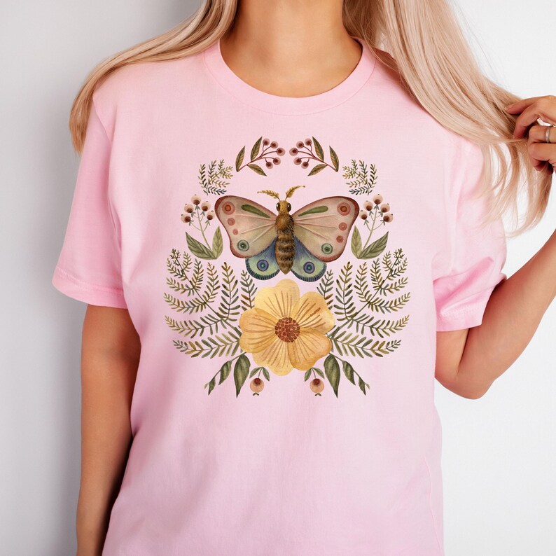 Pu&ograve; includere: T-shirt rosa chiaro con un disegno ad acquerello di una farfalla, un fiore giallo e fogliame. La farfalla ha ali beige, blu e rosa. Il fiore &egrave; giallo con un centro marrone. Il disegno &egrave; circondato da foglie e bacche verdi.