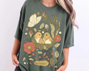 Könnte beinhalten: Ein olivgrünes Kurzarm-T-Shirt mit einem Grafikdruck von zwei Vögeln in einem Nest, umgeben von einem großen Schmetterling, orangefarbenen Blumen, grünen Blättern und einer einzelnen Feder. Die Illustration verwendet eine gedämpfte Farbpalette.